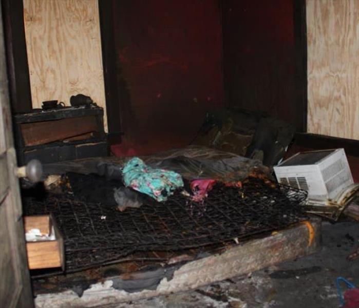 soot and smoke damaged matress, ac unit, and clothing in a boarded up bedroom