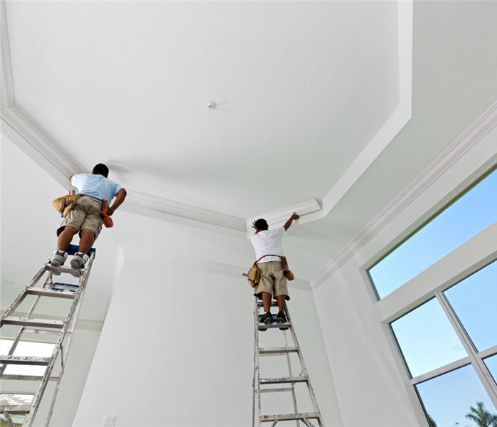Picture is of two men on ladders painting a commercial ceiling white. 
