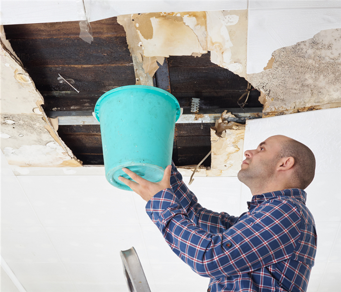 Picture is of a man holding a bucket to catch water coming out of a commercial paneled roof 